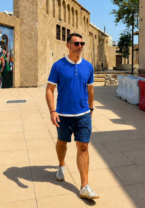 Man in a blue shirt and shorts walking on a sunny day with buildings and barriers in the background.