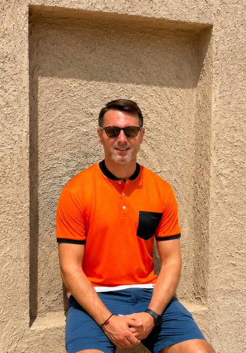 Man in an orange polo shirt and blue shorts sitting against a textured wall.