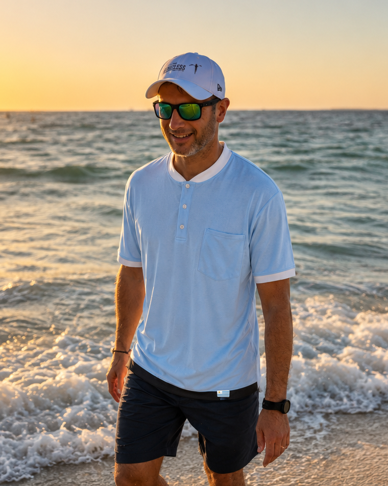 Man standing on a beach at sunset wearing a white cap, sunglasses, and a light blue shirt.
